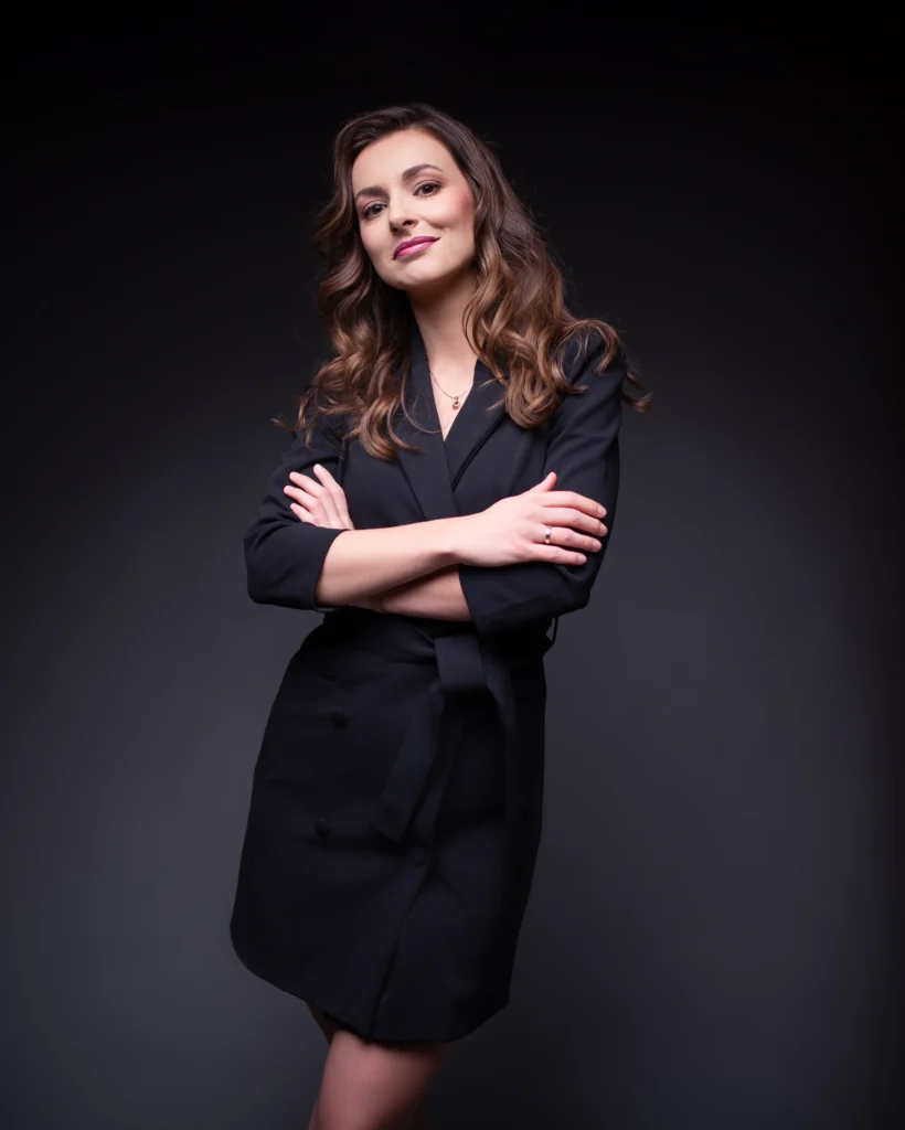 Portret Marceliny Machnik, radcy prawnego specjalizującego się w prawie budowlanym i nieruchomościach, uśmiechniętej, w czarnej sukience, na ciemnym tle